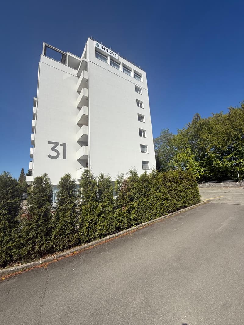 White building with multiple floors, labeled as '31', surrounded by hedges and trees, with a road in front