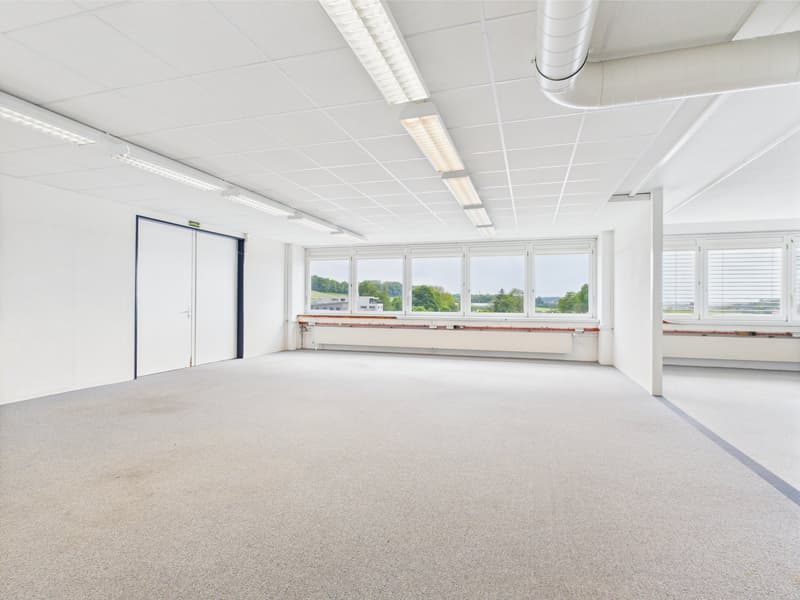 Spacious room, with carpeted flooring, recessed lighting, large windows with blinds, central door, and a radiator