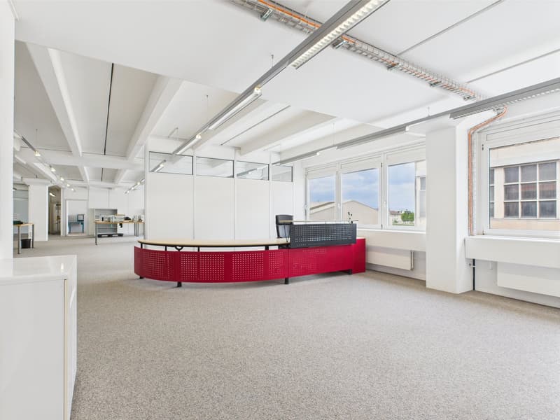 large open space, modern decor, red desk, white walls, large windows, fluorescent ceiling lights, large open desk, black chair