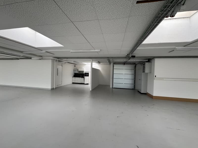 large open space, white walls, ceiling with grid and lights, concrete floor, garage and kitchen in the back