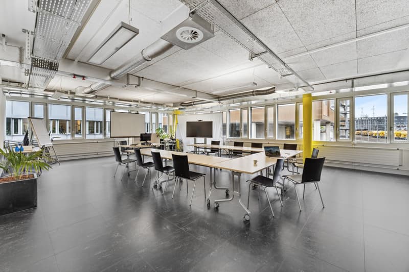 A spacious meeting room with glass windows, a projector screen, movable tables and chairs, and a whiteboard.