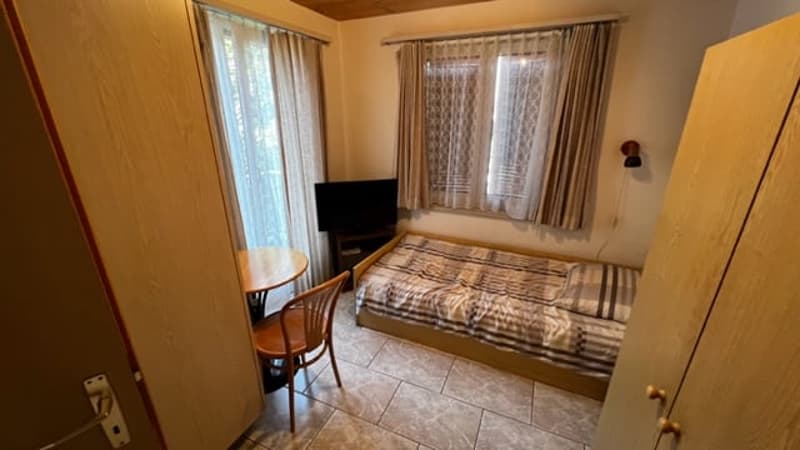 Bedroom with tile flooring, large windows, curtains, a bed with patterned bedding, a TV, and a small table with a chair.