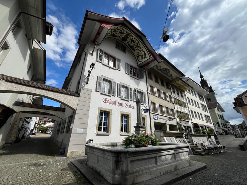The building has multiple floors with several windows. There is a fountain in front of the building with a flower arrangement. The building is made of white-colored wall and it has brown trim around the windows and archways. There is also a sign on the bu