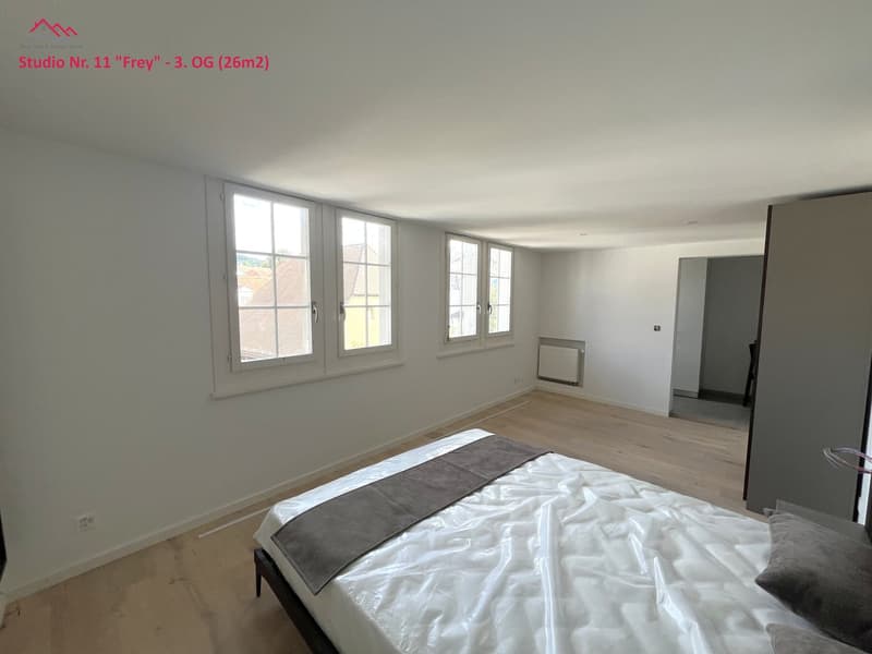 Empty bedroom with wooden floor and white walls, windows, bed with white mattress, wardrobe