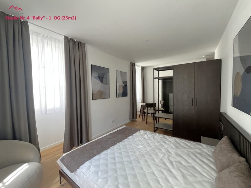 Bed, couch, hardwood floor, white walls, two picture frames, window with curtains, wooden chair and table, closet