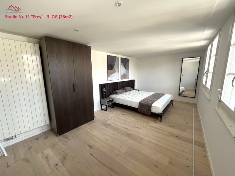 A room with a bed, a wardrobe, a nightstand, a mirror, and white windows.