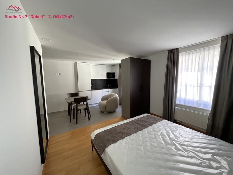 Bedroom with bed, chair, table, wardrobe, kitchen with microwave, sink, and cabinets, open windows, radiator, mirror.
