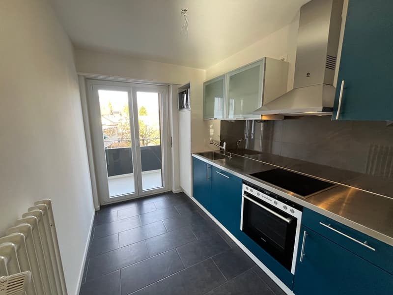 Modern kitchen with dark grey tiles, blue cabinets, stainless steel countertop, gas stove, built-in oven, modern hood, sink with faucet, glass door to balcony, radiators