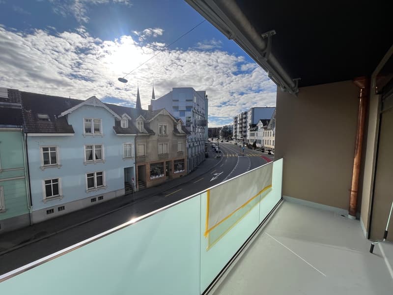 Balcony with glass railings, road with cars, buildings, streetlights, and cloudy sky