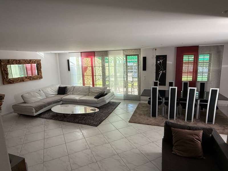 Spacious open-plan living room with large windows, gray sectional sofa, glass coffee table, and a dining area with a modern speaker system. The room has tiled flooring and access to a balcony or terrace.
