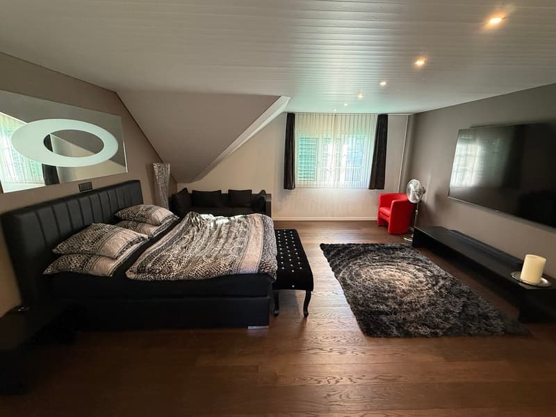 Spacious bedroom with hardwood flooring, large windows, a black leather bed frame, patterned bedding, a red accent chair, and a shag rug.