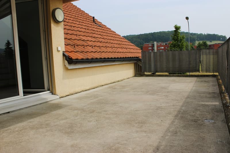 The image shows a balcony or terrace area of an apartment. The balcony has a tiled roof and a concrete floor. There is a window visible on the building, and the surrounding area includes some trees and other buildings in the background.