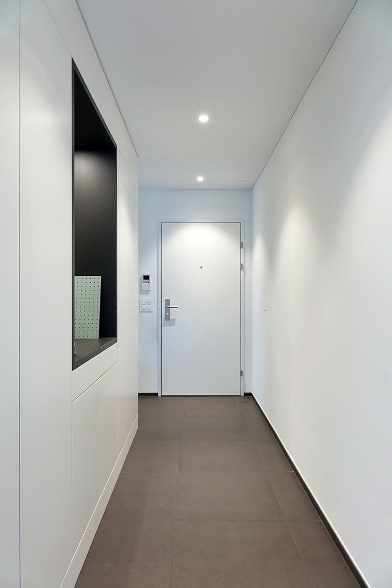 White walls, brown tiled floor, closed door, small shelf, hallway