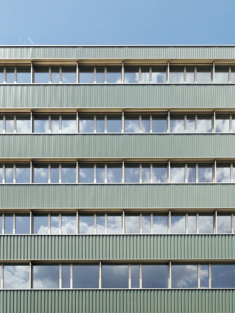 Multi-story building with a uniform facade of green metal panels and large windows. The building appears to be a commercial or office structure.