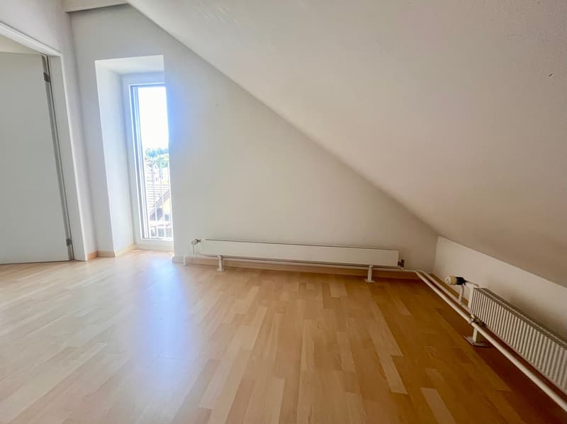 Spacious bedroom with hardwood flooring, slanted ceiling, and a window providing natural light. The room features a radiator heating system.