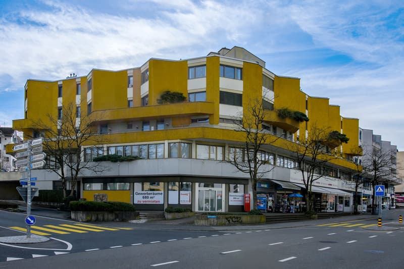 A yellow and white multi-story building with glass windows and balconies, located on a street with shops and trees.