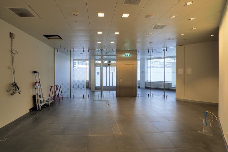 large empty room, glass walls, tiled floor, ceiling lights, a ladder and some tools are on the side of the room