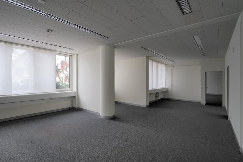 Empty room with carpeted floor, white walls, windows with white curtains, central column, ventilation grilles on the ceiling.