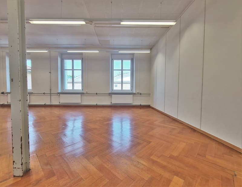 empty room with wooden parquet flooring, three windows, ceiling mounted lamps, white walls, central heating radiators