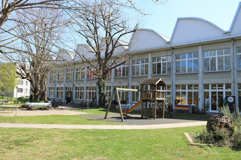 Modern building with glass windows, a playground with a slide and a swing, greenery, benches and tables