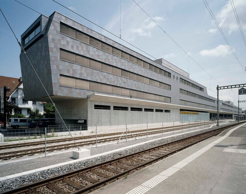 A modern, multi-story building with a sleek, geometric facade made of concrete and glass. The building has a rectangular shape with multiple floors and large windows. It is located next to a railway track, with the building's entrance and ground floor vis