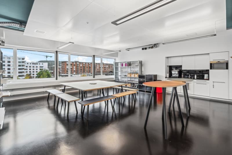 Large open office space with floor-to-ceiling windows providing a view of the surrounding buildings. The room has a modern, minimalist design with white walls, black and wood furniture, and a kitchen area with appliances.