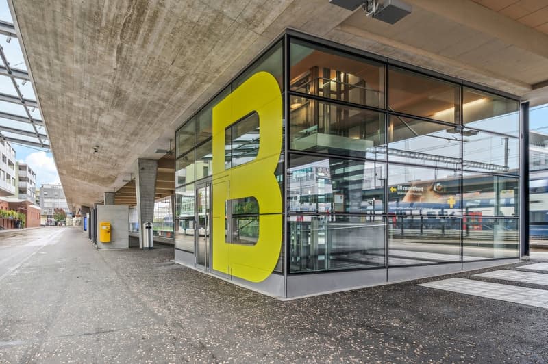 The image shows the exterior of a modern commercial building with a large yellow letter 'B' displayed prominently on the glass facade. The building has a minimalist, industrial design with concrete ceilings and a glass entryway. The surrounding area appea