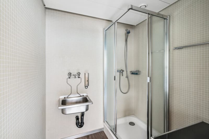 The image shows a modern bathroom with a glass-enclosed shower stall, a wall-mounted sink, and tiled walls in a light color scheme.