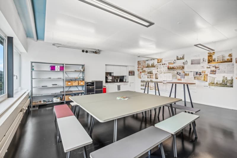 Large open workspace with a long table, benches, and shelving units. The walls are decorated with various images and posters. There is a kitchen area with appliances visible, including a dishwasher.