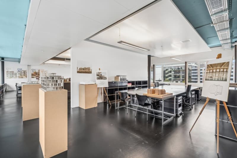 Large open-plan office space with high ceilings, white walls, and concrete flooring. The space includes desks, chairs, and various architectural models and displays. There are also some storage boxes and an easel visible, indicating this is likely a desig