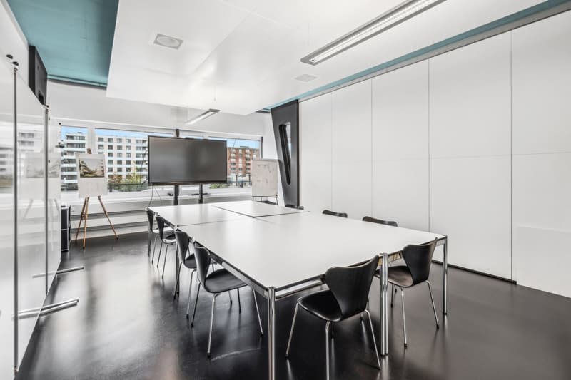 Large conference room with a long white table, black chairs, a mounted TV screen, and large windows providing a view of the surrounding buildings.