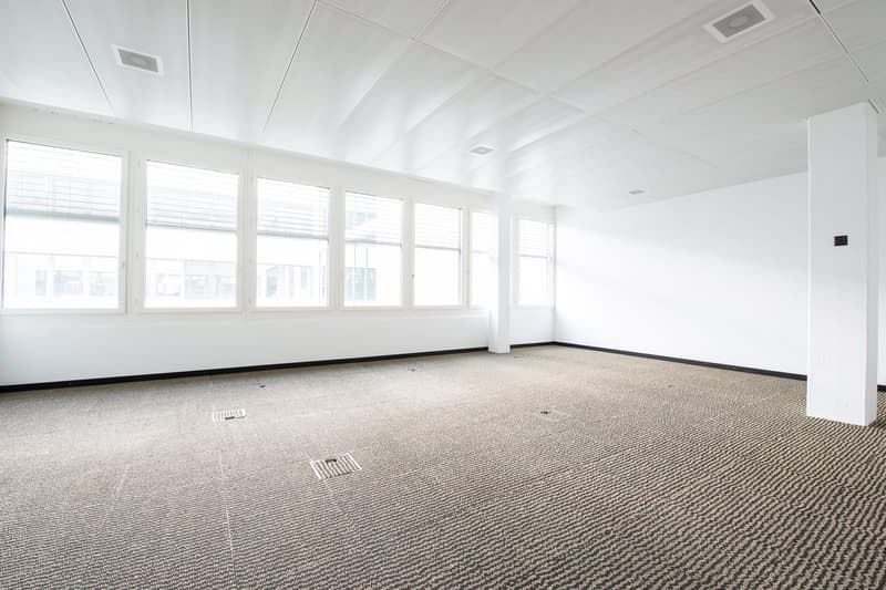Large open office space with white walls, large windows, and a carpeted floor. The room appears to be empty and ready for use.