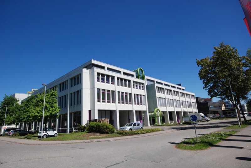 Two-story office building, white exterior, large windows, glass doors, multiple parked cars in front, shrubs, streetlights, trees, and a sign