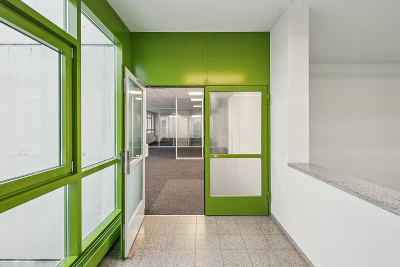 A narrow corridor with white tiled flooring, green walls, and large glass doors.