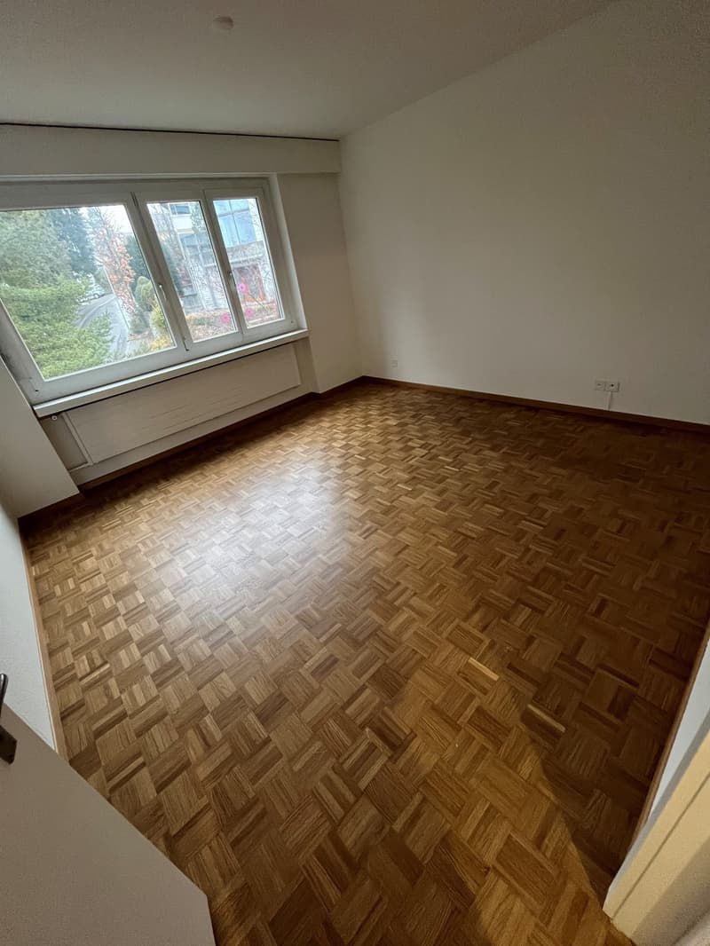 empty room with wooden parquet flooring, large window with outdoor view, white walls, two power outlets