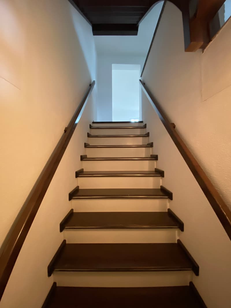 The image shows a staircase with wooden steps and a white wall on either side. The staircase appears to be leading up to a bright, open space at the top.