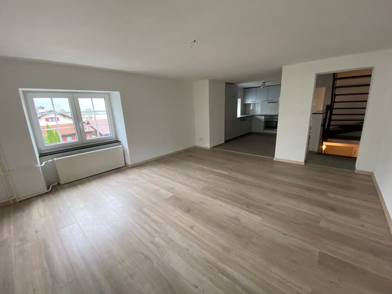 The image shows a spacious open-plan living area with a large window, hardwood flooring, and a modern kitchen visible in the background. The room appears to be newly renovated with a clean, minimalist design.