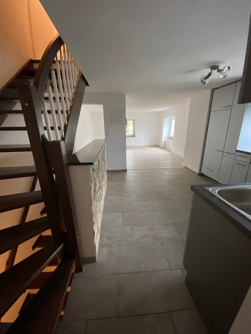 The image shows the interior of a residential property. It features a wooden staircase leading up to the second floor, with a tiled floor in the entryway and open-plan living space visible beyond. The space appears to be newly renovated, with clean white