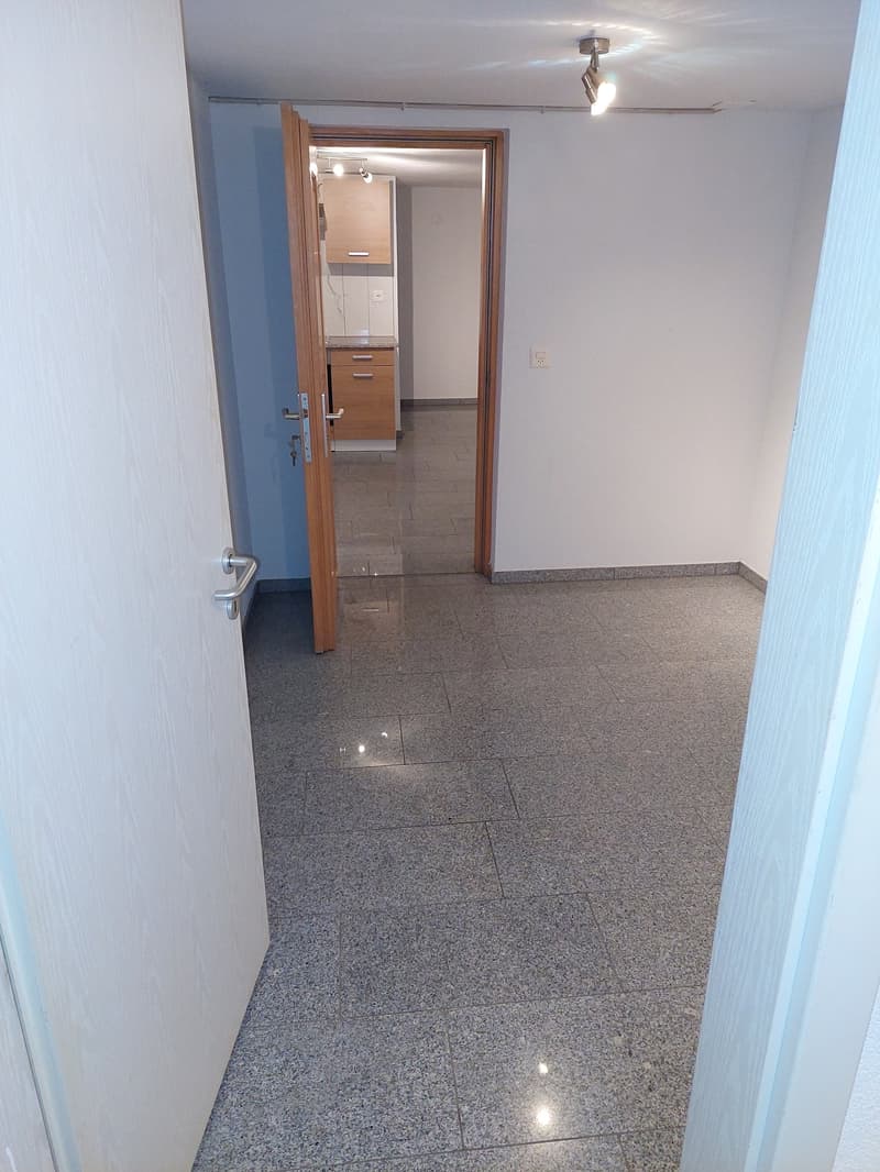 The image shows the interior of an apartment. The room has white walls and a granite tile floor. There is a wooden door frame leading to another room, which appears to be a kitchen or bathroom. The lighting in the room is provided by a ceiling-mounted spo