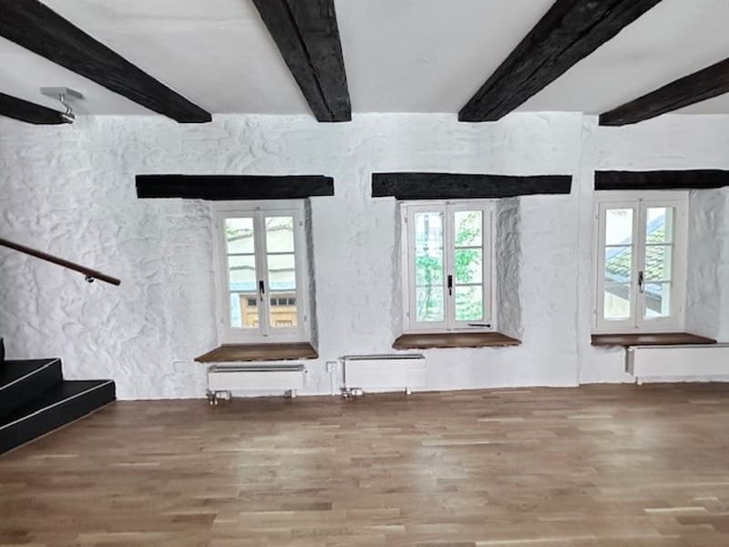 large open space with wooden flooring, white walls, exposed wooden beams, three windows with wooden frames, built-in bench seating under the windows, radiators and power outlets installed on the wall