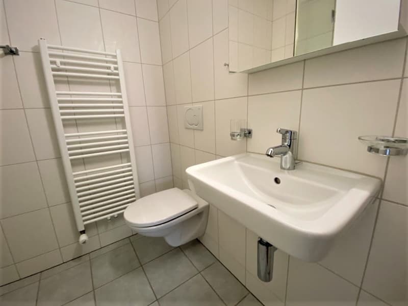 A small bathroom with white tiles, a toilet, a sink, and a towel rack.