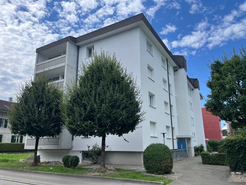 White apartment building with multiple floors, balconies, and windows, surrounded by trees and bushes, with a driveway and a red building in the background.