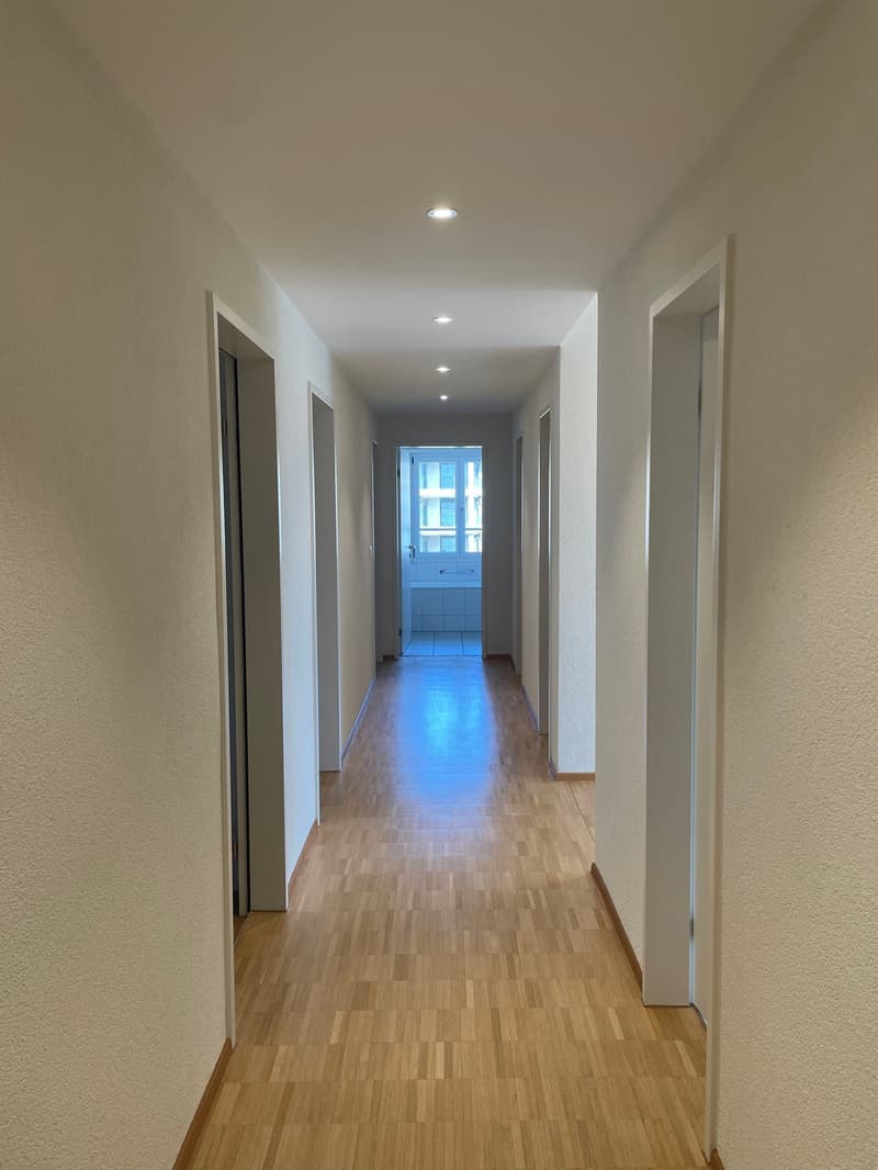 Empty hallway with wooden floors, white walls, and ceiling lights.