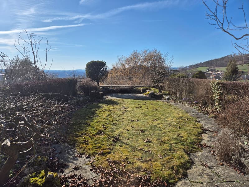A lawn with several patches of grass, surrounded by trees and shrubs, a paved walkway, and a distant view of a village and mountains.