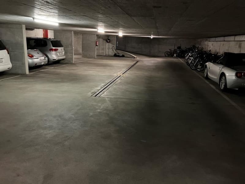 Underground parking garage with concrete walls and floor, several cars and motorcycles parked, lighting fixtures on the ceiling