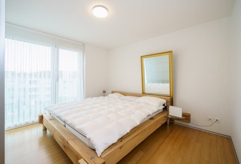 wooden bed, white duvet, white pillows, mirror, lamp, wooden floor, white walls, large windows