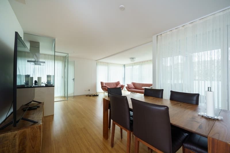 Dining room with wooden floor, large windows, dining table, chairs, TV, and couches