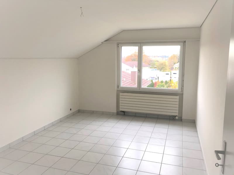 Spacious attic room with large windows, tiled floor, and radiator. The room has a bright and airy feel with a view of the surrounding neighborhood.