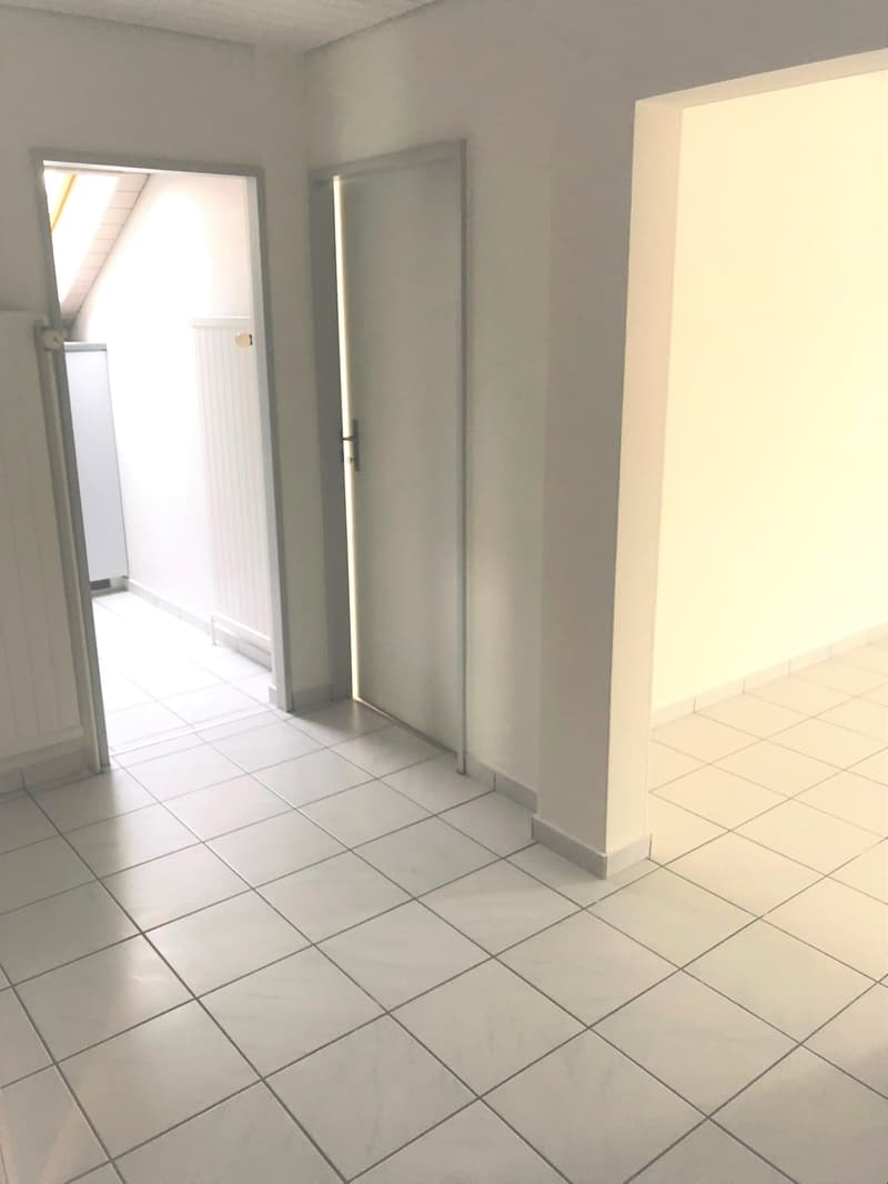 The image shows an empty room with white walls and gray tile flooring. There is a door visible in the background, and a mirror on the wall to the right.