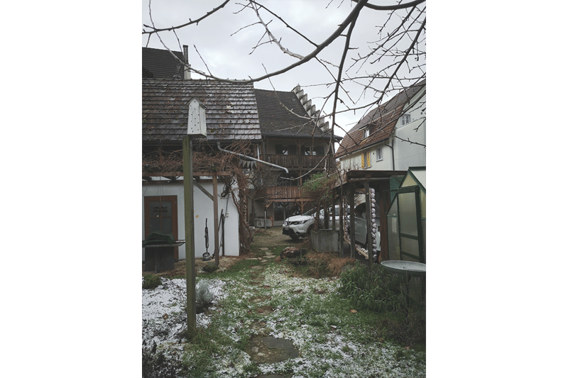 Three houses next to each other with snowy ground. Vehicles parked behind a house and a walkway. Trees without leaves in front of the houses.